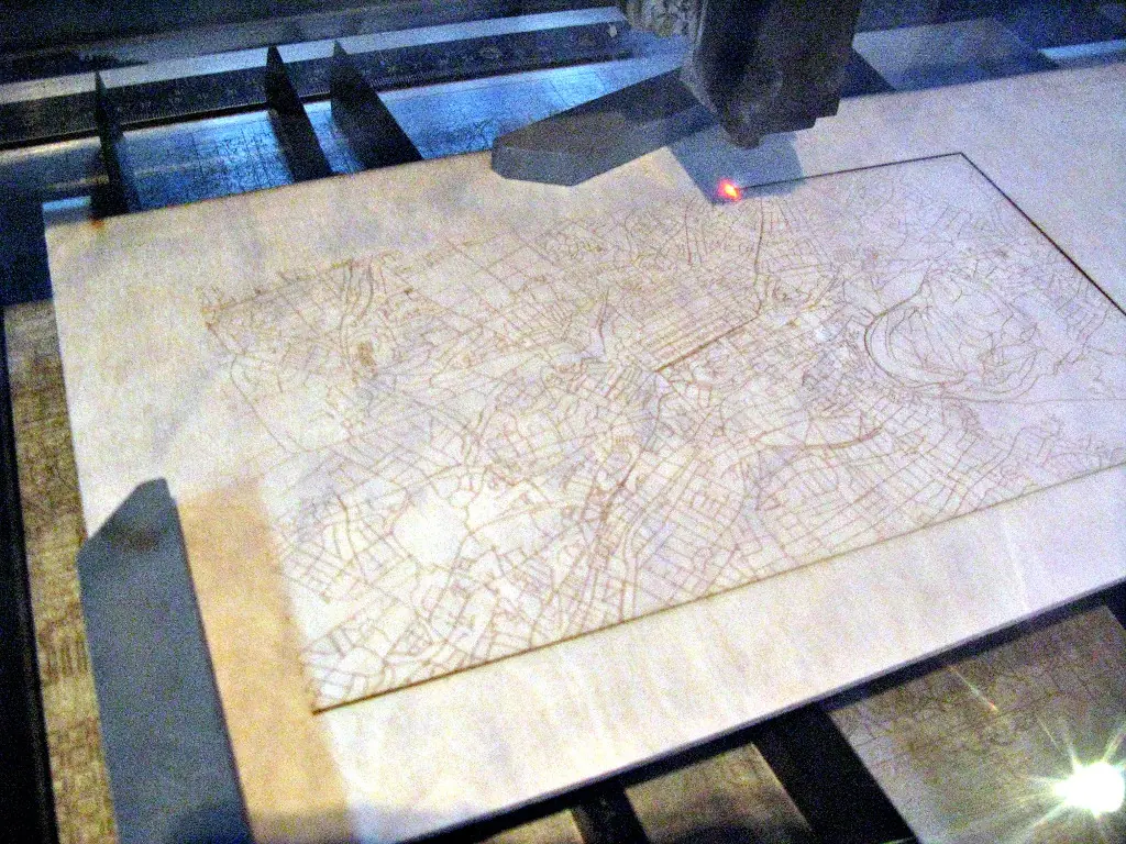 A laser cutter burning through a plywood sheet engraved with a street map, seen from above with the head over the sheet and a bright point where the beam meets the wood