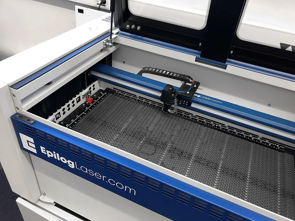 The open bed of a desktop CO2 laser: honeycomb bed, rulers along the edges, a gantry carrying the lens head on a drag chain