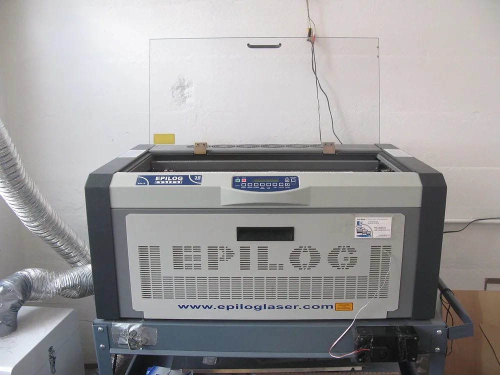 A desktop CO2 laser cutter on a stand in a workshop, with a flexible exhaust duct running from its left side up to the wall