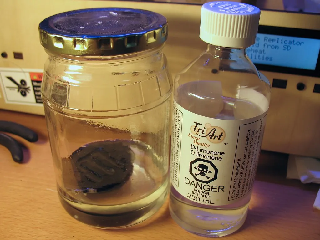 A dark printed part soaking in a glass jar of clear liquid next to a bottle of d-limonene with a hazard label