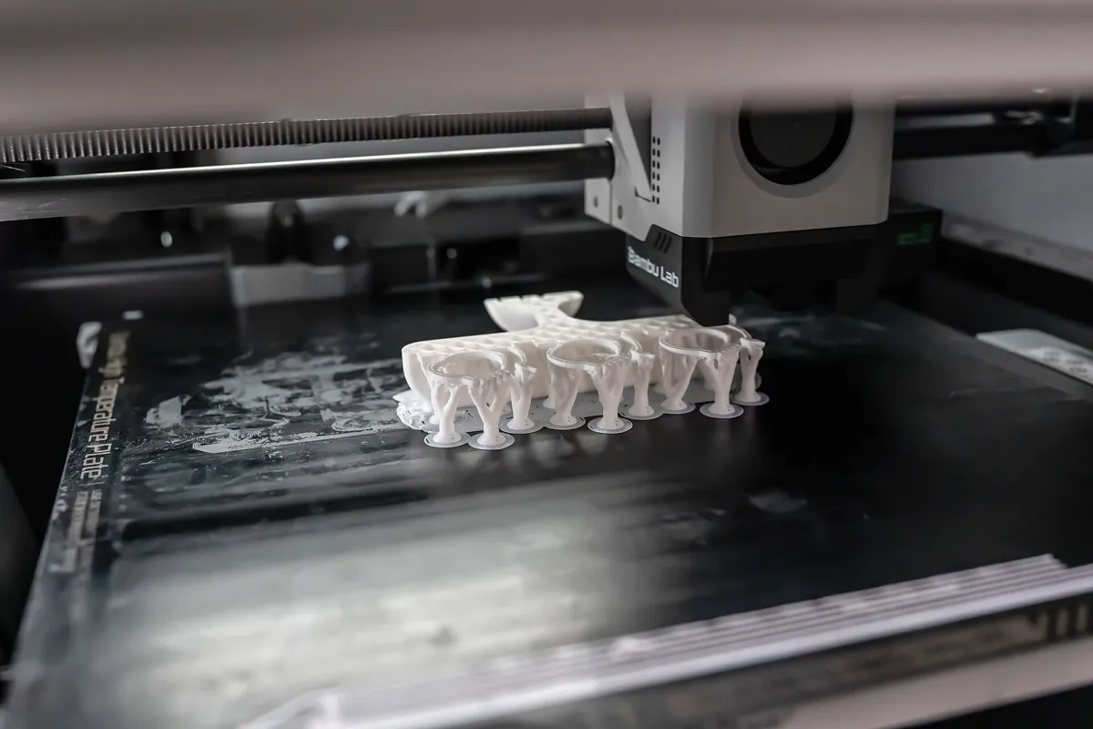 Inside a Bambu Lab X1 Carbon: the toolhead prints a white part that rests on a row of thin support trunks, each standing on a small round foot on the build plate